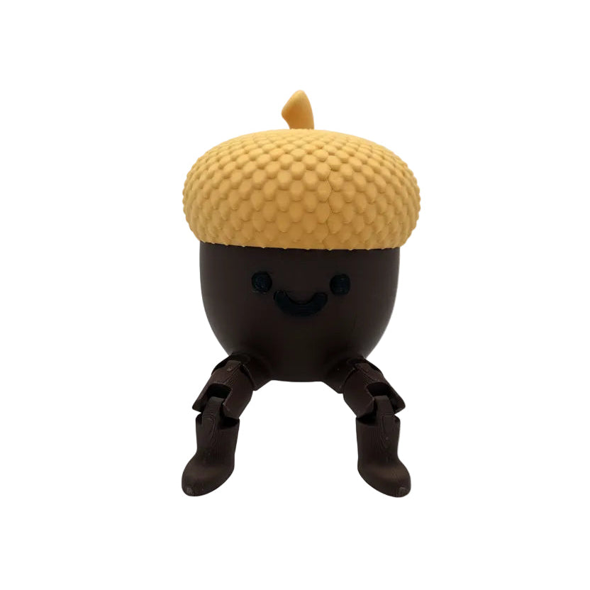 A small acorn-shaped figurine with a smiling face, sitting on a clear acrylic block against a light gray studio background. The acorn has a textured golden-yellow cap and dark brown body and legs.