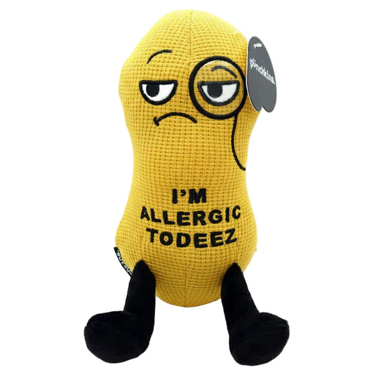 A yellow crocheted plush toy shaped like a peanut with black legs and a sad face, featuring the text 'I'M ALLERGIC TO DEEZ' and a small side tag.