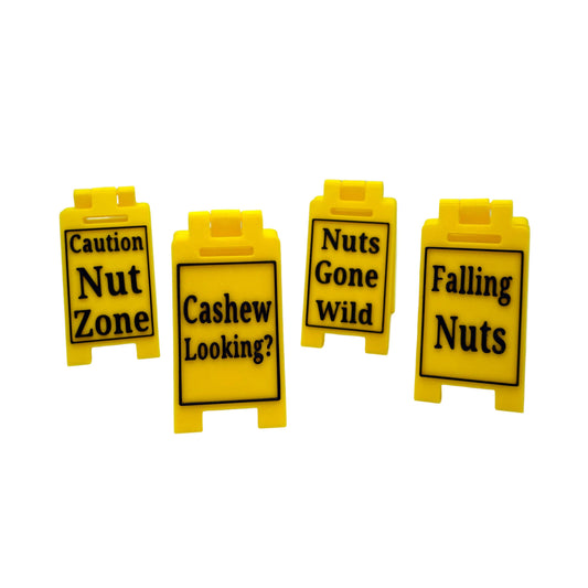 Four small yellow plastic sign-shaped items with black text reading 'Caution Nut Zone,' 'Cashew Looking?,' 'Nuts Gone Wild,' and 'Falling Nuts' on a light gray studio background.