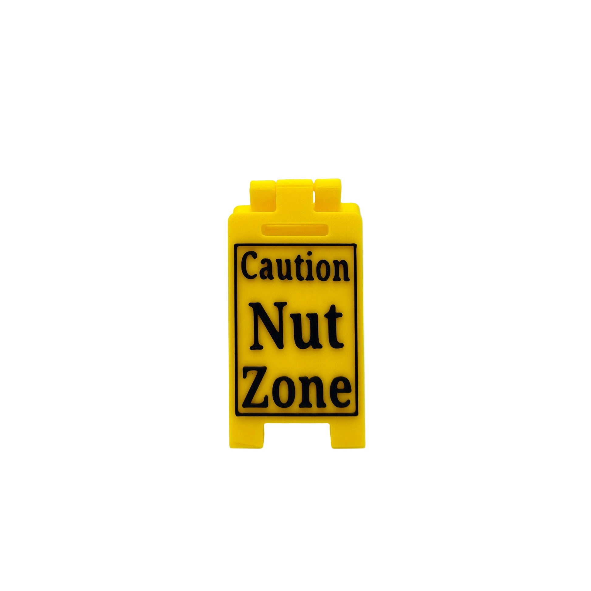 A small yellow plastic sign with black text reading 'Caution Nut Zone' on a white studio background.