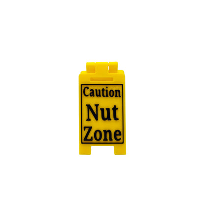 A small yellow plastic sign with black text reading 'Caution Nut Zone' on a white studio background.