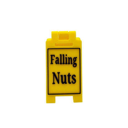 A small yellow plastic sign with black text reading 'Falling Nuts' on a white studio background.