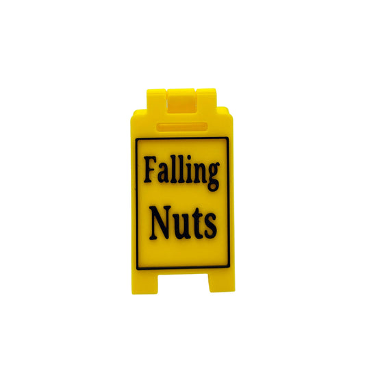 A small yellow plastic sign with black text reading 'Falling Nuts' on a white studio background.