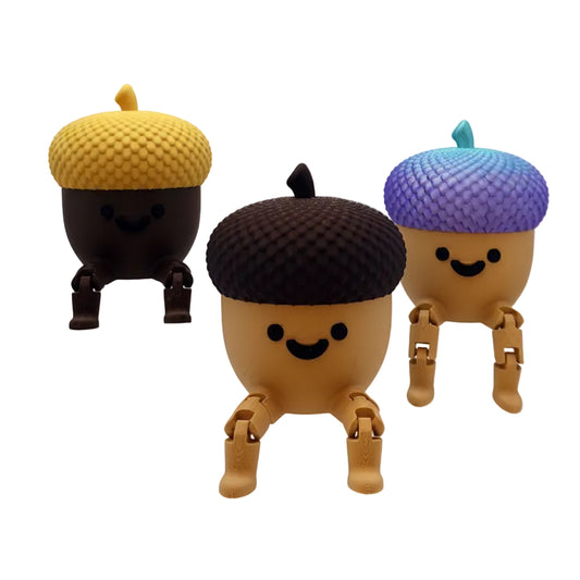 Three cute acorn-shaped figurines with smiling faces, displayed on clear acrylic stands against a white background. The acorns have textured caps in yellow, dark brown, and purple-blue, with tan bodies and legs.
